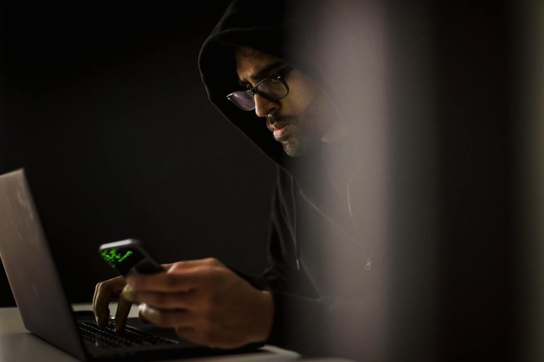Crop bearded ethnic male computer geek in hoodie chatting on cellphone while typing on netbook in dark room